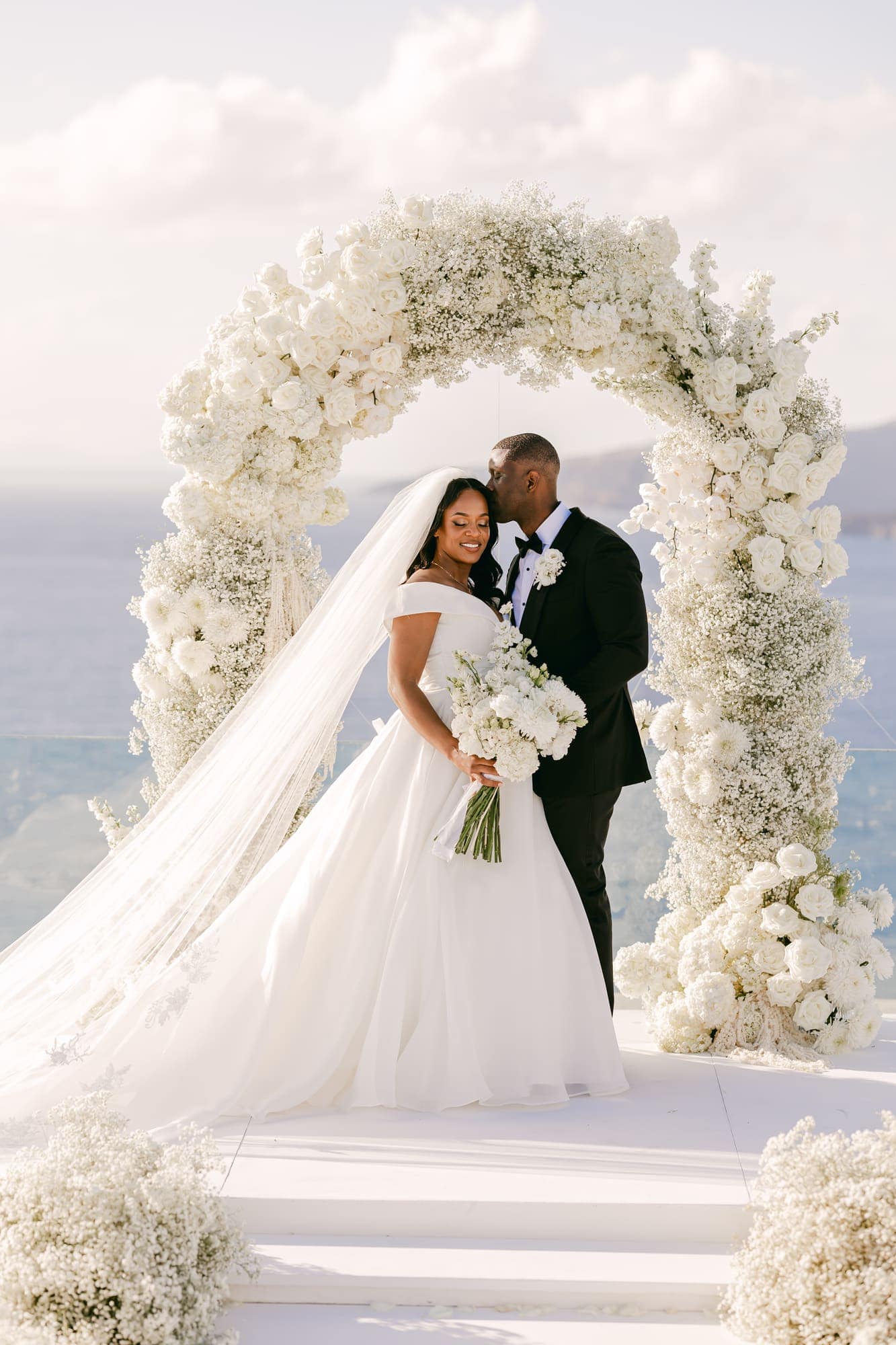 Irlon & Jailleen — portraits beneath the arch of white roses and baby's breath, Caldera terrace, Santorini
